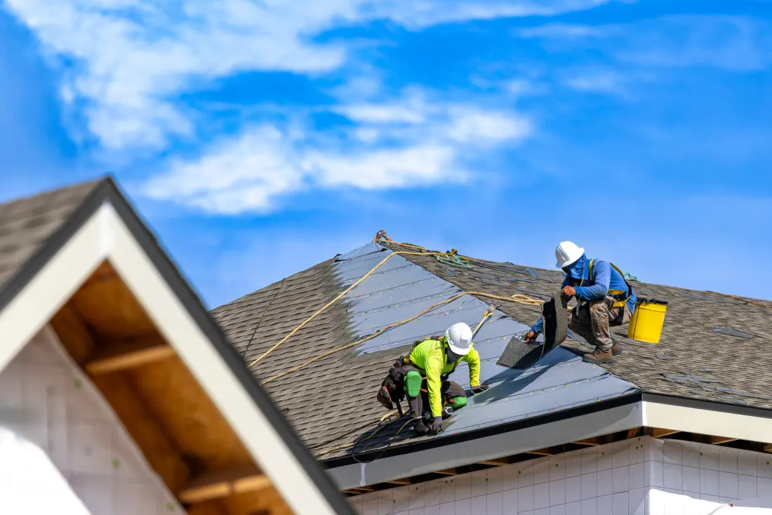 Two roofers installing new asphalt shingles on a house under a clear sky