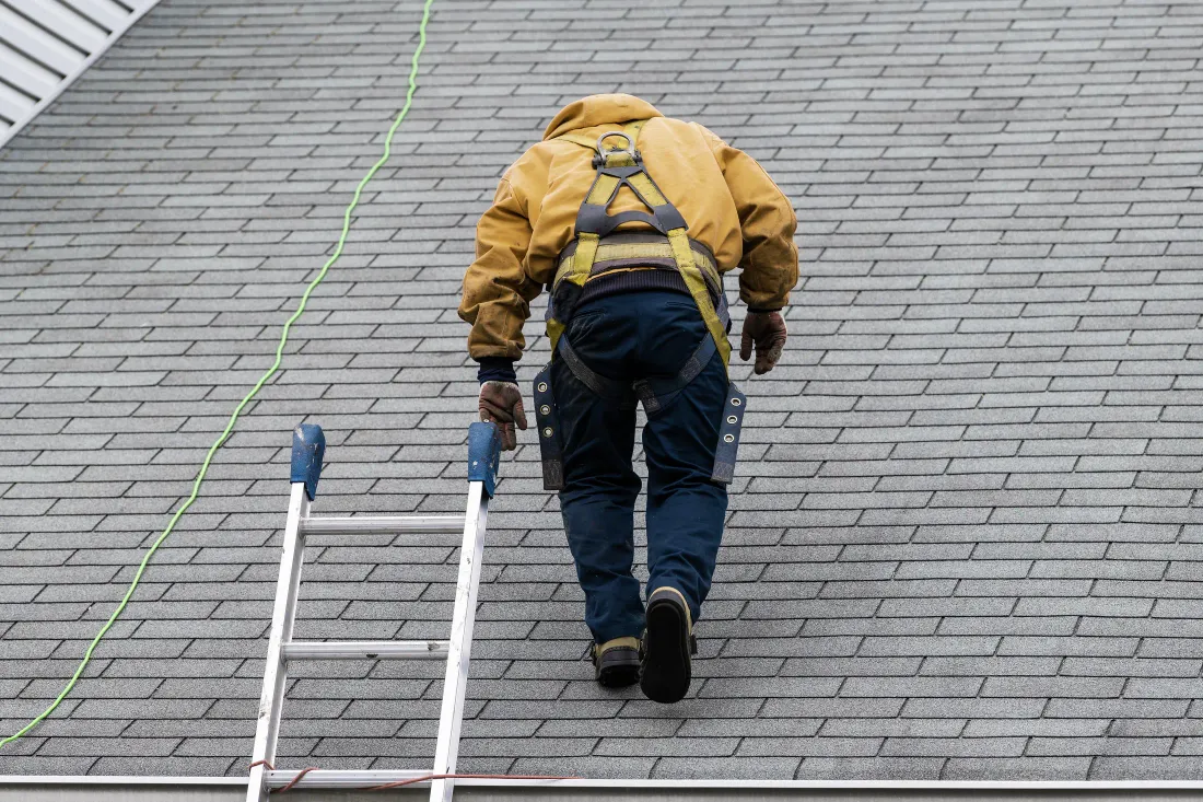 A roofer in a safety harness walking a shingle roof near a ladder during a roof inspection