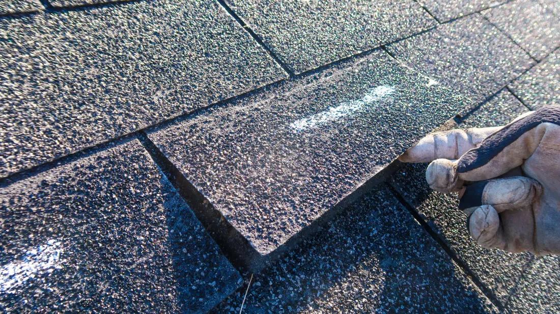 A gloved hand checking an asphalt shingle for repair, close up